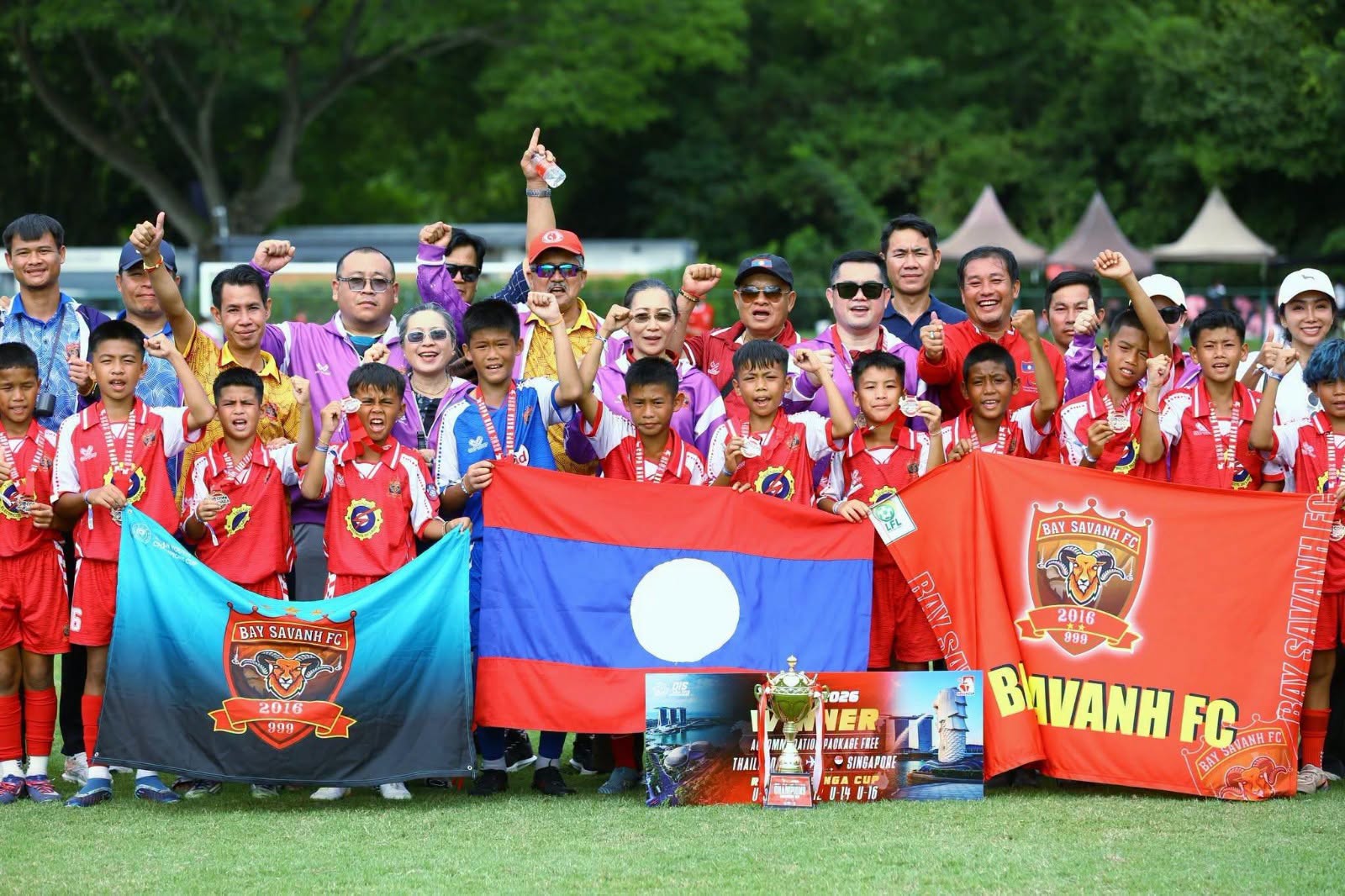 Baysavanh FC under-10 players and officials celebrate with the Lao flag, club banners and the COPA NAKA 2026 trophy